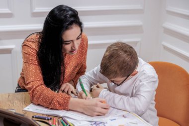 Koyu renk saçlı bir eğitimci, gözlüklü sarışın bir çocuğa ödevini zarif bir ortamda yapmasına yardım eder..