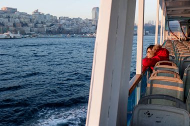 Kırmızı ceketli kız feribotla karşıya geçerken uzaklara bakıyor.