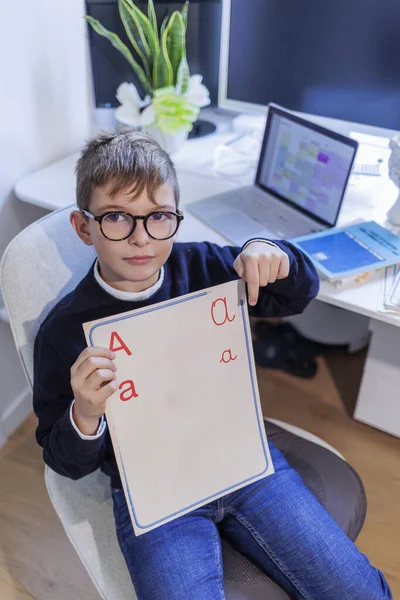 金发男孩戴着眼镜 身穿蓝色衣服 坐在桌子前 他看到一块标有字母字母的板子 桌上有计算机和监视器 — 图库照片