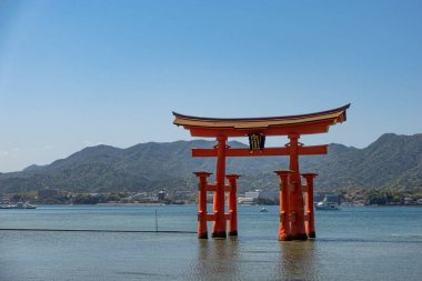 Canlı bir kırmızı torii kapısı zarif bir su kütlesinin ortasında duruyor. Arka planda heybetli dağlar yükseliyor.