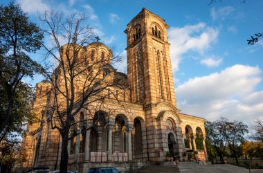 Büyük bir saat kulesi barındıran büyük, etkileyici bir tuğla bina güzel bir şekilde yemyeşil ağaçlar ve tepemizde parlak mavi bir gökyüzü ile çevrilidir.