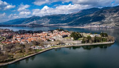 Geniş bir hava manzarası, uzak arkaplanda görkemli dağlarla çevrili sakin bir gölün tam ortasında bulunan küçük, sakin bir adayı gözler önüne seriyor.