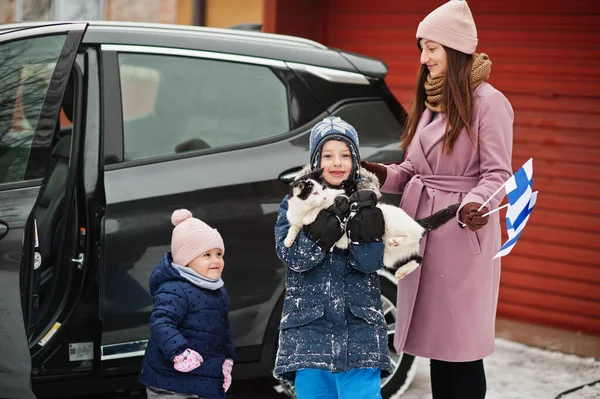 stock image Kids with cat on hands against car in the yard of house at winter. Mother hold flag of Finland.