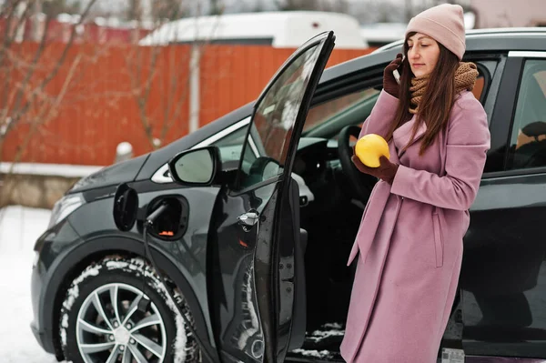 Young woman with mobile phone and citrus lemon fruit at hand charging her electric car in winter. Eco transport concept.