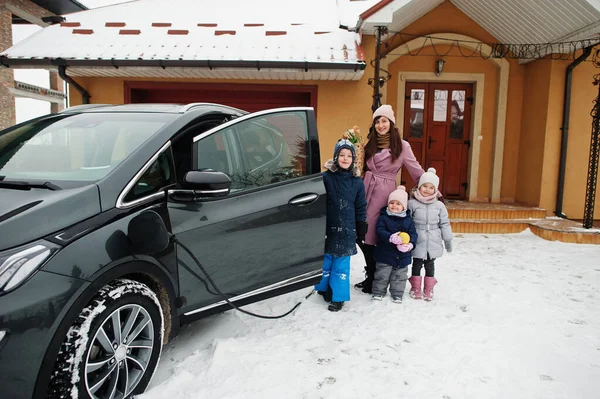 Young woman with kids hold eco bags and charging electric car in the yard of her house .
