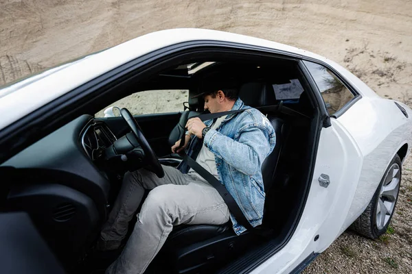 Handsome man in jeans jacket and cap sit and tie belt at his white muscle car.