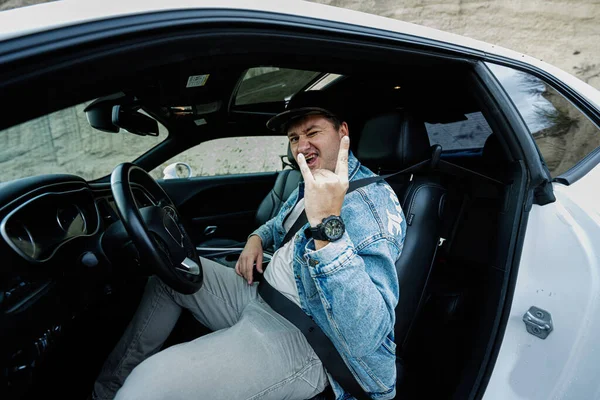 Handsome man in jeans jacket and cap sit at his white muscle car and show Rock and Roll hand sign.