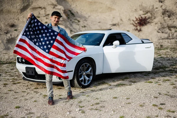 Handsome man in jeans jacket and cap with USA flag near his white american muscle car in career.
