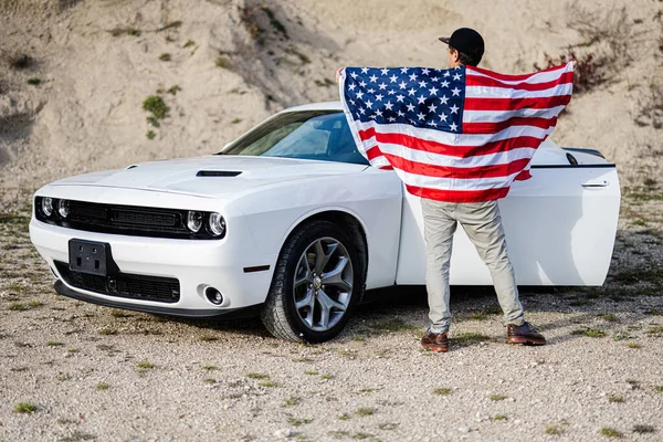 Back of man with USA flag near his white american muscle car in career.