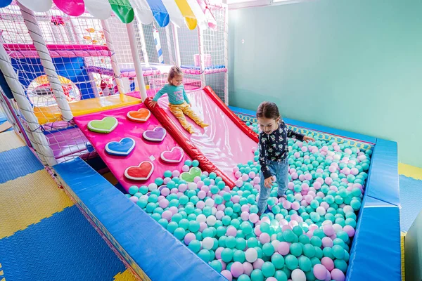 Happy sisters playing at indoor play center playground, color balls in ball pool.