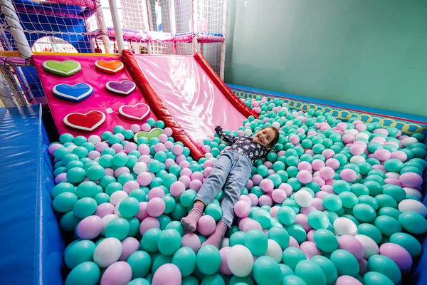 Happy girl playing at indoor play center playground. Lying at color balls in ball pool.