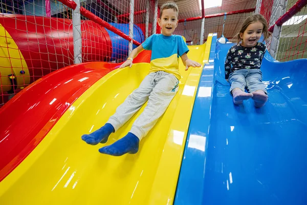 Brother with sister playing at indoor play center playground. Children slides in colored slide.