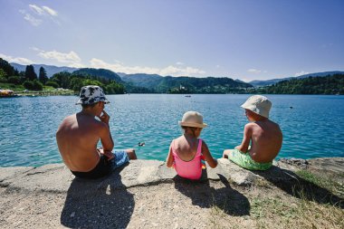 Panamalı üç çocuk manzaralı iskelede oturuyor Güzel Bled Gölü, Slovenya.