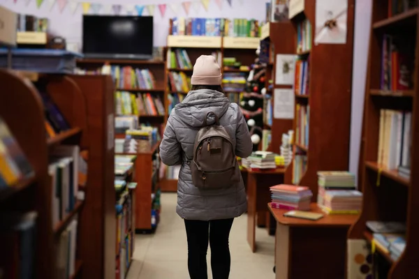 Back of woman in jacket and backpack reaching a book from bookshelf at the library.