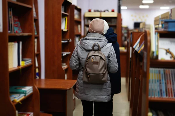 Back of woman in jacket and backpack reaching a book from bookshelf at the library.