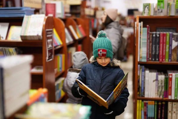 Boy in jacket and hat reaching a book from bookshelf at the library. Learning and education of european kids.