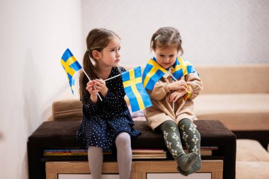 Two sisters are sitting on a couch at home with swedish flags on hands. Sweden children girls with flag .