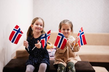 Two sisters are sitting on a couch at home with norwegian flags on hands. Norway children girls with flag .