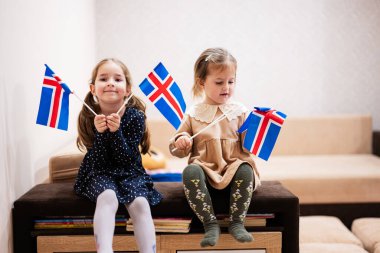 Two sisters are sitting on a couch at home with icelandic flags on hands. Iceland children girls with flag .