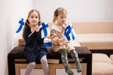Two sisters are sitting on a couch at home with finnish flags on hands. Finland children girls with flag .