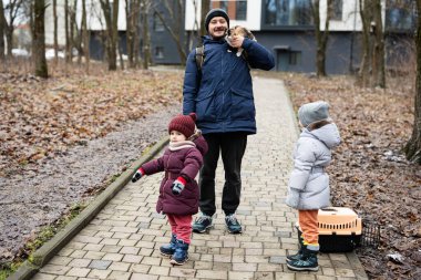 Father and daughters with cat in travel pet plastic cage carriage outdoor at park.