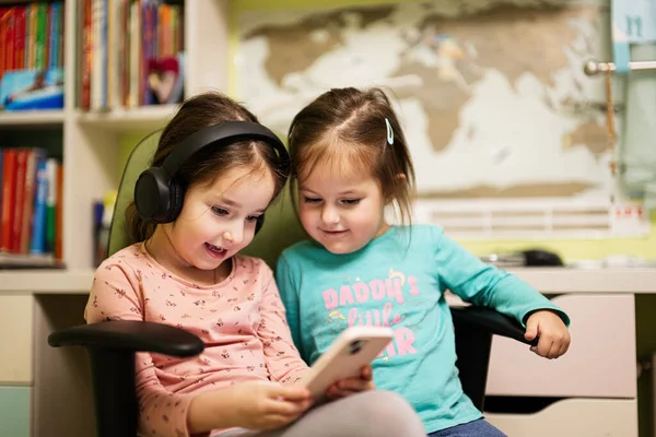 Two little girls sisters wear headphones watching cartoons or kid video on mobile phone.