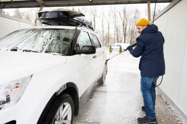 Man washing high pressure water american SUV car with roof rack at self service wash in cold weather.