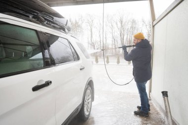 Man washing high pressure water american SUV car with roof rack at self service wash in cold weather.