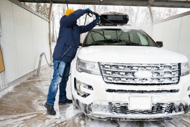 Man washing high pressure water american SUV car with roof rack at self service wash in cold weather.