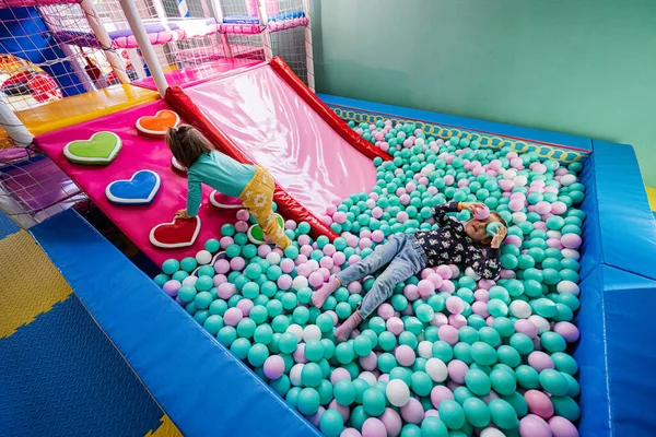 Happy sisters playing at indoor play center playground. Girl lying at color balls in ball pool.