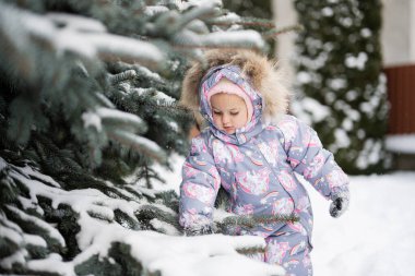 Baby girl play outdoors in snow near Christmas tree in winter. 
