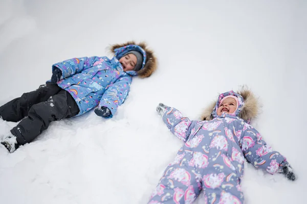 Two sisters making snow angel while lying on snow.