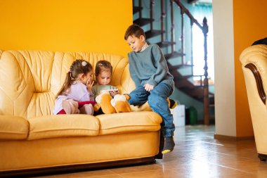 Children sit on a leather sofa in the living room and watch cartoons on phone.