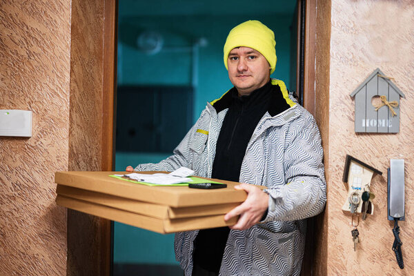 Delivery man with pizza cardboard boxes. Courier in green color hat standing against door of residential house