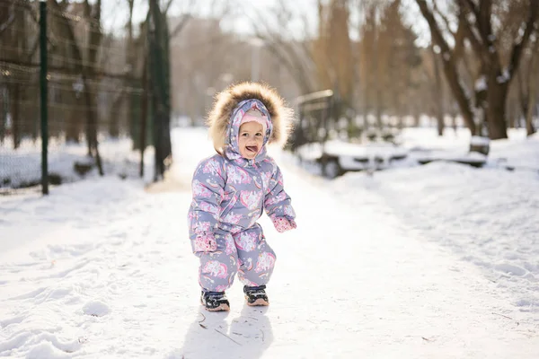 Baby girl show tongue, wear child snowsuit on a sunny frosty winter day.