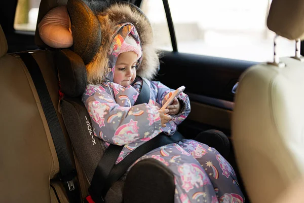Child safety seat chair baby girl with mobile phone at hands is on back seat of car.