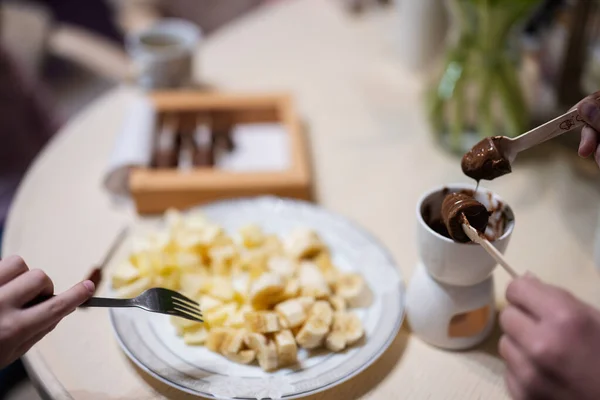 Children eat fruits and desserts, drink tea at home in the evening kitchen. Close up hand with chocolate on a stick for melting.