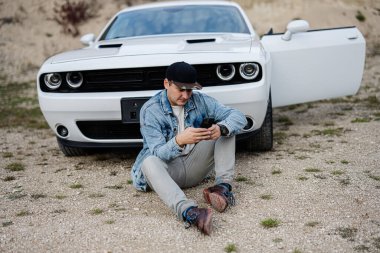 Handsome man in jeans jacket and cap sitting near his white muscle car in career and texting on phone.