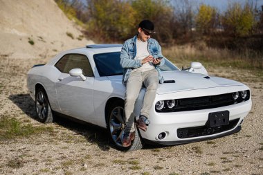 Handsome man in jeans jacket and cap sit on hood at his white muscle car in career and texting on phone.