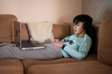 Young teen boy with headphones in front of a laptop on a bed at evening.
