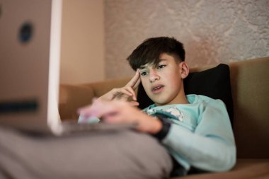 Young teen boy in front of a laptop on a bed at evening.