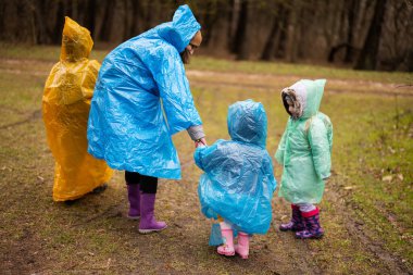 Rear view of mother and three children walking in the forest after rain in raincoats together.