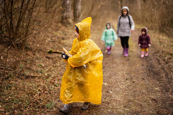 Boy in yellow raincoat with log at hands, against mother and children in the forest after rain in raincoats together.