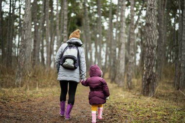 Back of mom with backpack and daughter walking along the forest after rain together.