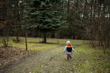 Back of baby girl with backpack run along the forest after rain.