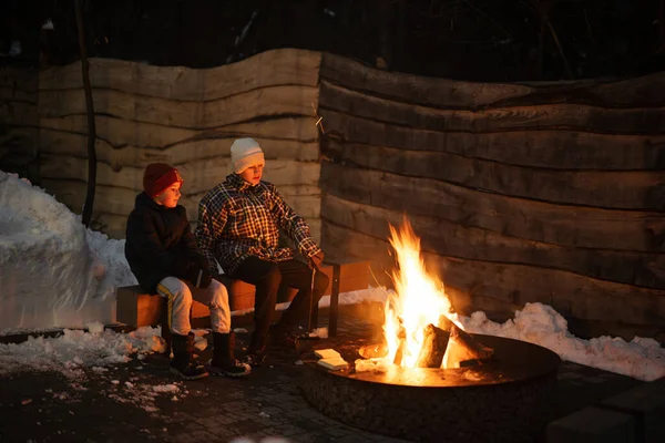 Two brothers sit by the fire pit in winter night.