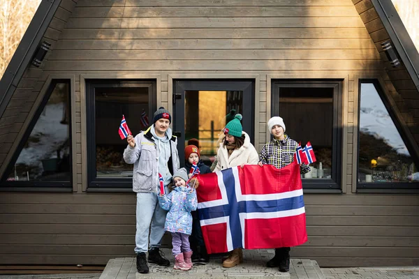 Portrait of family with kids outside cabin house holding Norway flags. Scandinavian culture, norwegian people.