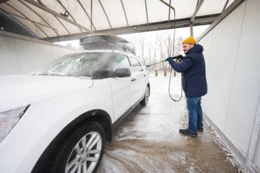 Man washing high pressure water american SUV car with roof rack at self service wash in cold weather.