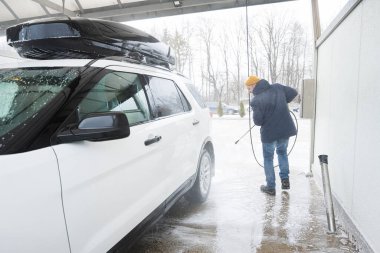 Man washing high pressure water american SUV car with roof rack at self service wash in cold weather.