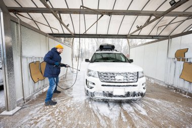 Man washing high pressure water american SUV car with roof rack at self service wash in cold weather.